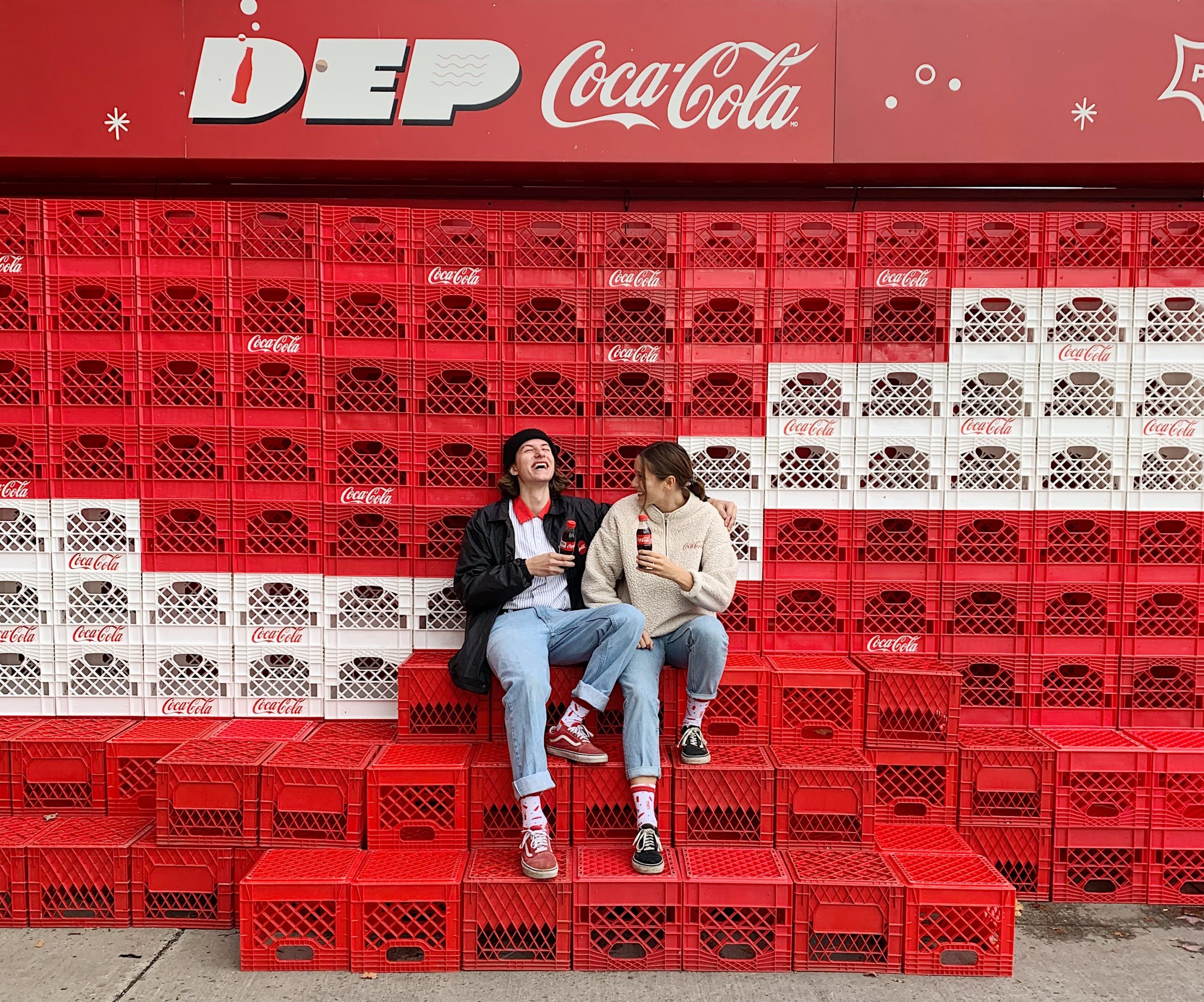 Two people sitting on milk crates under a Coca Cola sign