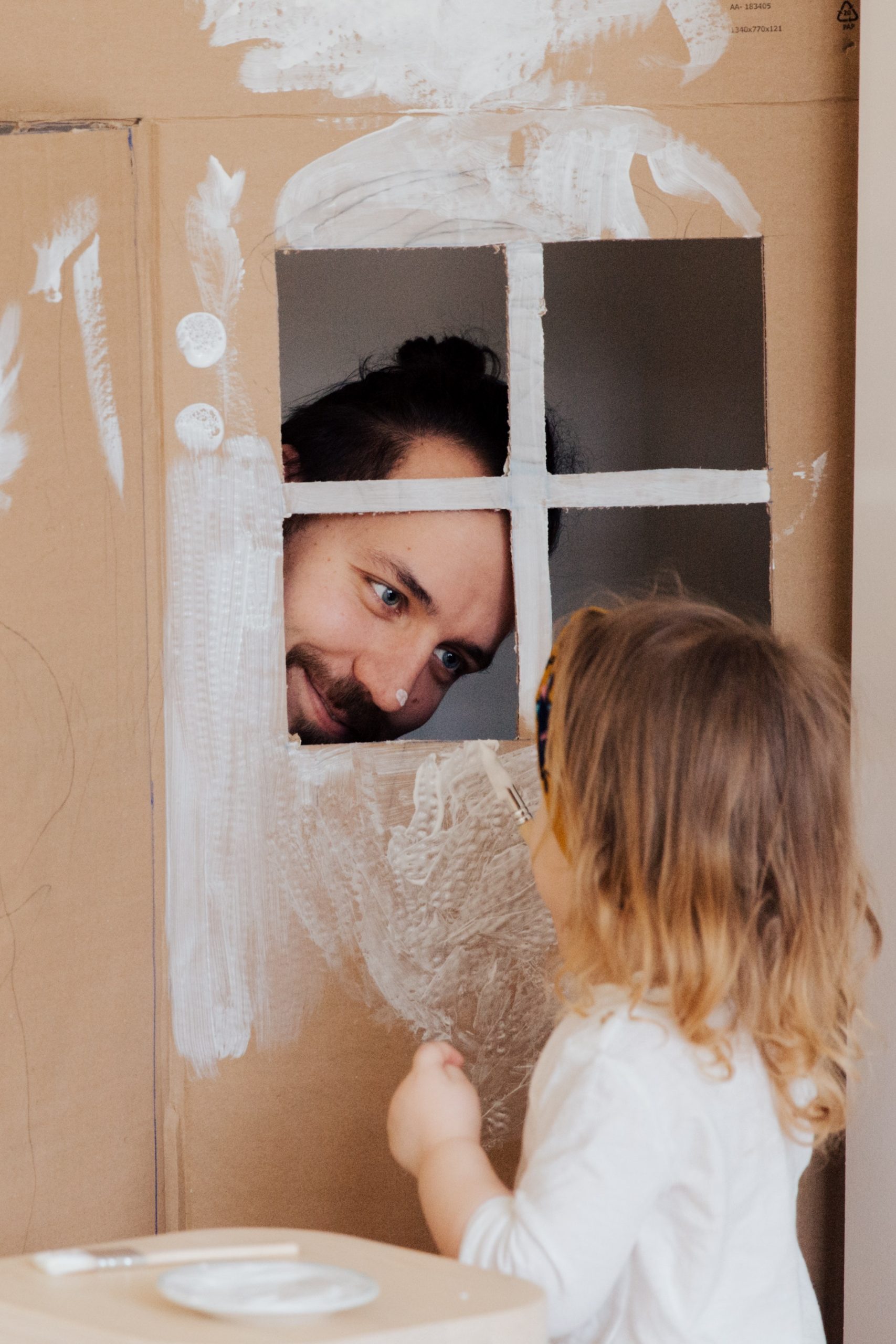Father in cardboard playhouse with daughter, white paint.